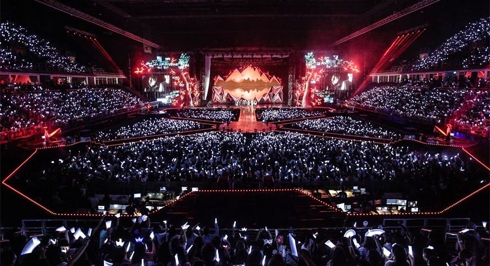 High-angle shot of a live performance in a packed stadium with a vibrant light show and a sea of glowing lightsticks in the crowd.