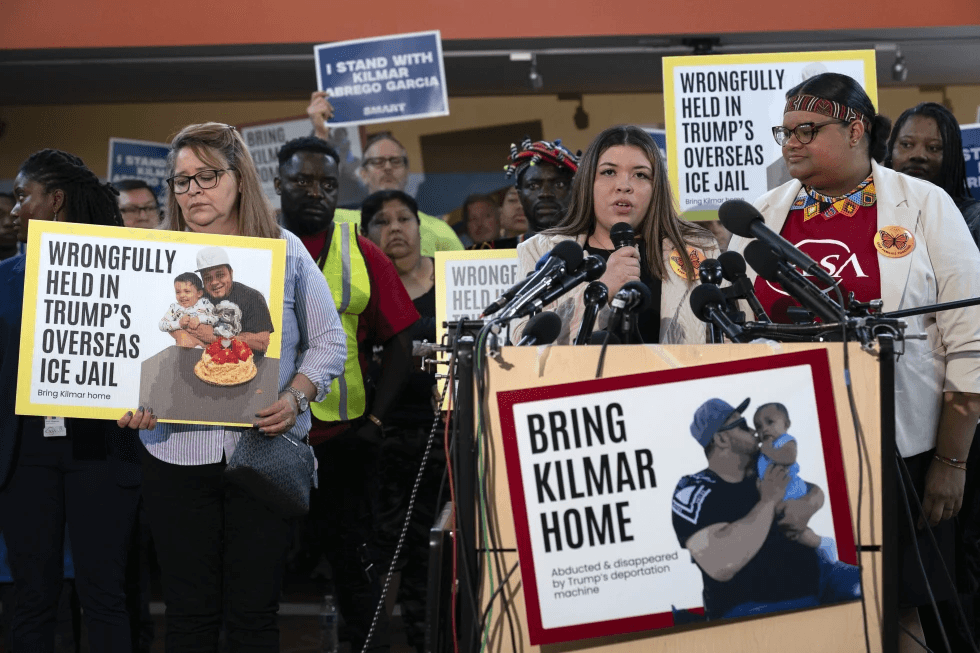 Jennifer Vasquez Sura speaking at a news conference with signs reading "WRONGFULLY HELD IN TRUMP'S OVERSEAS ICE JAIL" and "BRING KILMAR HOME".